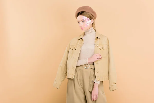 Jolie femme avec des fleurs sur le visage portant béret et debout isolé sur beige — Photo de stock