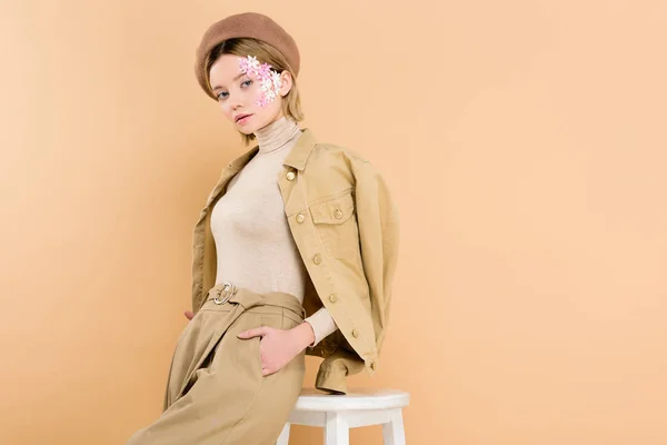 Trendiges Mädchen in Baskenmütze steht mit der Hand in der Tasche in der Nähe von Stuhl isoliert auf beige — Stockfoto