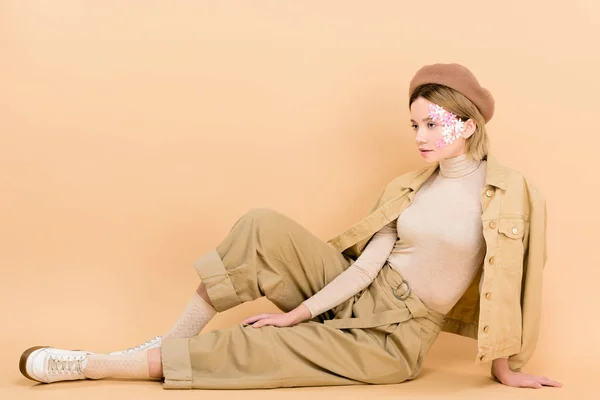 Stylish girl in beret posing while sitting on floor isolated on beige — Stock Photo