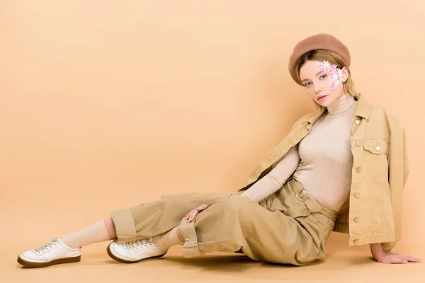 Trendy girl in beret posing while sitting on floor isolated on beige — Stock Photo