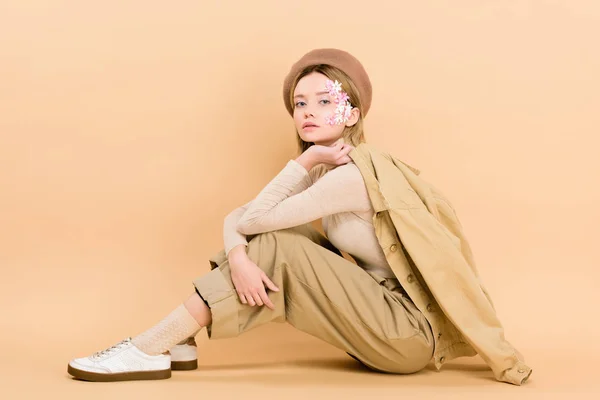 Chica atractiva en boina posando mientras está sentado en el suelo aislado en beige - foto de stock