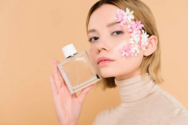 Beautiful woman with flowers on face holding bottle of perfume isolated on beige — Stock Photo