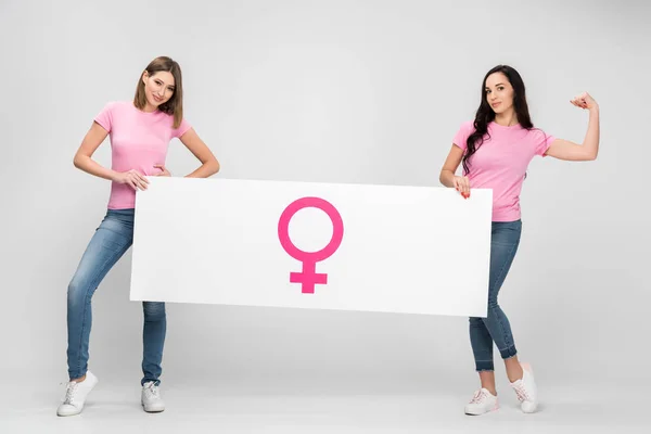 Beautiful woman showing muscle and holding large sign with female symbol with friend on grey background — Stock Photo