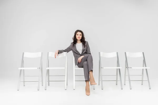 Attraktive Frau sitzt mit überkreuzten Beinen auf Stuhl isoliert auf grau — Stockfoto