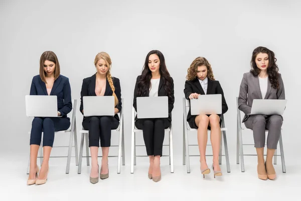 Attraktive junge Frauen mit Laptops, während sie auf Stühlen sitzen, die isoliert auf grau sind — Stockfoto