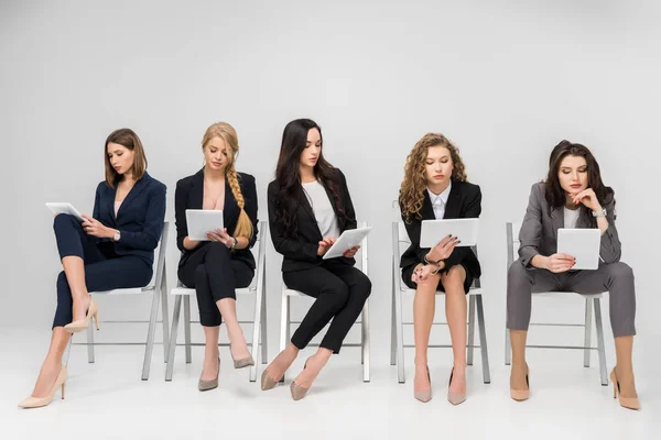 Séduisantes femmes d'affaires utilisant des tablettes numériques tout en étant assis sur des chaises isolées sur gris — Photo de stock
