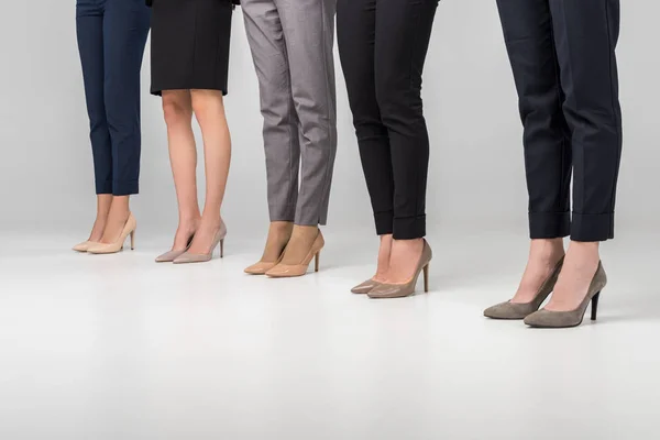 Vista recortada de las mujeres de pie en zapatos de tacón alto sobre fondo gris - foto de stock