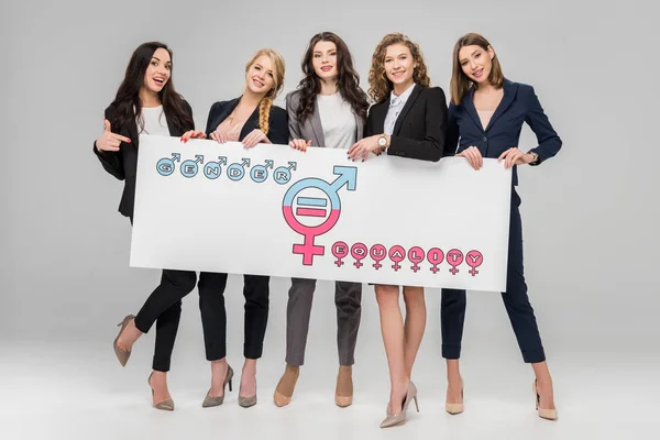 Alegres mujeres jóvenes sosteniendo un gran cartel con símbolo de igualdad de género sobre fondo gris - foto de stock