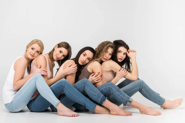 Hübsche junge Frauen, die einander die Köpfe auf die Schultern legen, während sie auf grauem Hintergrund sitzen und sich umarmen — Stockfoto
