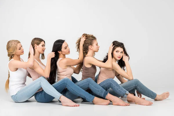 Hermosas chicas tocando el pelo de la otra mientras se sientan juntos sobre fondo gris - foto de stock