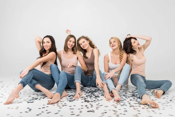 Веселые девушки, сидящие вместе и смеющиеся рядом со звездами конфетти, изолированными от серых — стоковое фото