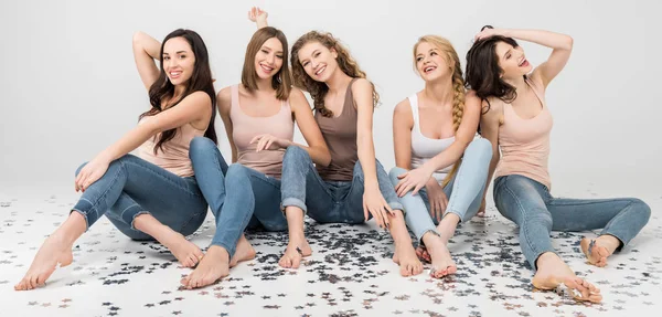 Счастливые девушки, сидящие вместе и смеющиеся рядом со звездами конфетти, изолированными от серых — стоковое фото