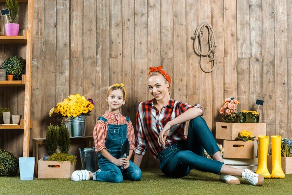 Cheerful woman sitting on grass with daughter near wooden fence — Stock Photo