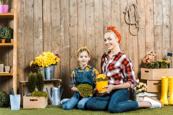 Daughter sitting with cheerful mother on grass and holding plants near wooden fence — Stock Photo