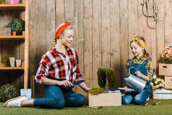 Madre che tiene pala vicino figlia impianto irrigazione mentre seduto su erba vicino recinzione di legno — Foto stock