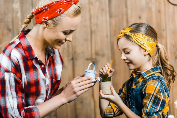 Счастливая мать поливая маленькое растение с игрушечной лейкой банка рядом с милой дочерью — стоковое фото