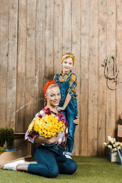 Cheerful woman holding flowers and sitting near daughter on grass — Stock Photo