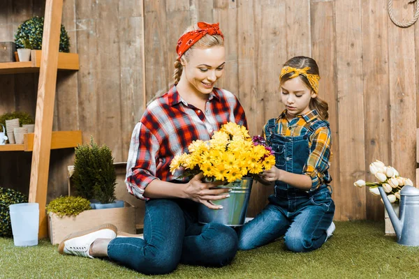 Cheerful woman holding flowers and sitting near cute daughter on grass — Stock Photo