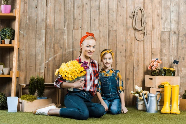 Mujer feliz sosteniendo flores y sentado con linda hija en la hierba - foto de stock