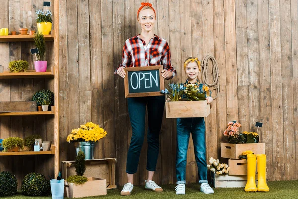 Attractive woman holding small chalkboard with open lettering near cute daughter holding box with plants — Stock Photo