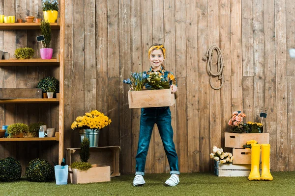 Cute kid holding box with plants and standing near wooden fence — Stock Photo