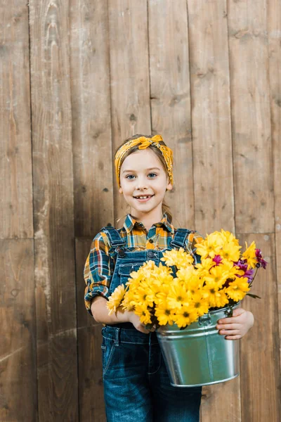 Enfant heureux tenant des fleurs dans un seau et debout près d'une clôture en bois — Photo de stock