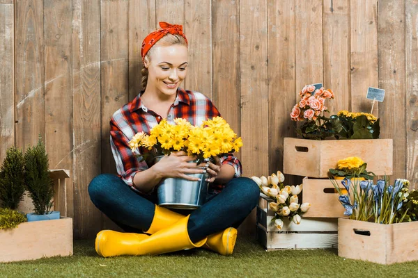 Glückliche Frau, die mit überkreuzten Beinen sitzt und Blumen im Eimer betrachtet — Stockfoto