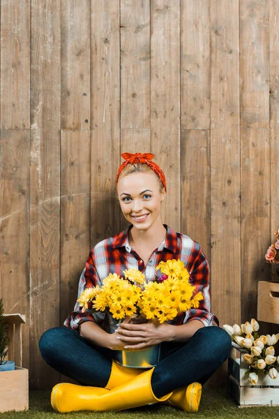 Mujer feliz sentada con las piernas cruzadas y sosteniendo flores en cubo - foto de stock