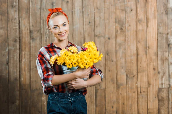 Bela mulher segurando flores em balde e sorrindo perto de cerca de madeira — Fotografia de Stock