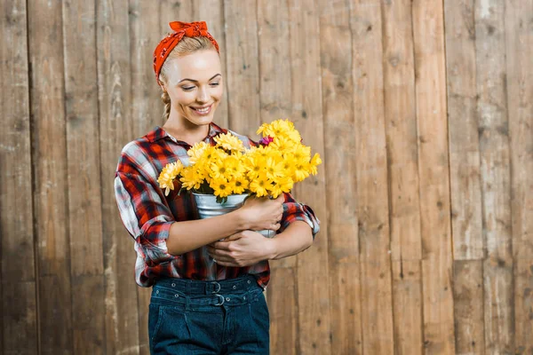Beautiful woman looking at flowers in bucket and smiling near wooden fence — Stock Photo
