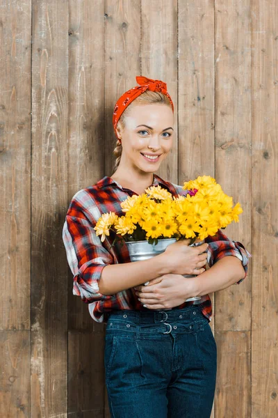 Mulher feliz segurando flores em balde e sorrindo perto de cerca de madeira — Fotografia de Stock