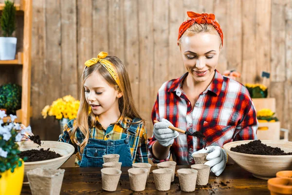 Schöne Mutter hält kleine Schaufel und legt Boden in leeren Pappbecher in der Nähe niedliche Tochter — Stockfoto