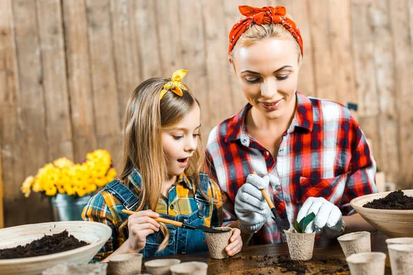 Alegre madre sosteniendo pequeña pala cerca de pequeña planta en taza de papel cerca de hija sorprendida - foto de stock