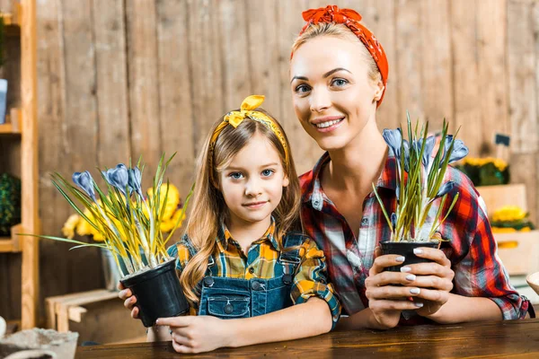 Cheerful mother and cute daughter looking at camera while holding pots with flowers — Stock Photo