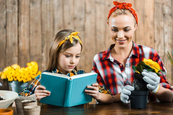 Joyeuse mère tenant des fleurs avec des reflets près de pot et en regardant fille livre de lecture — Photo de stock