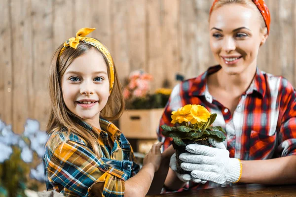 Selektiver Fokus des fröhlichen Kindes, das in die Kamera neben der Mutter blickt, die einen Topf mit Blumen hält — Stockfoto