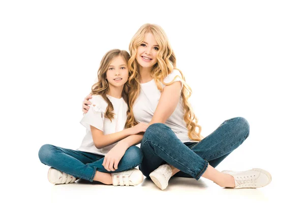 Mutter und Tochter umarmen sich, während sie mit überkreuzten Beinen auf dem Boden sitzen und in die Kamera auf weißem Hintergrund schauen — Stockfoto