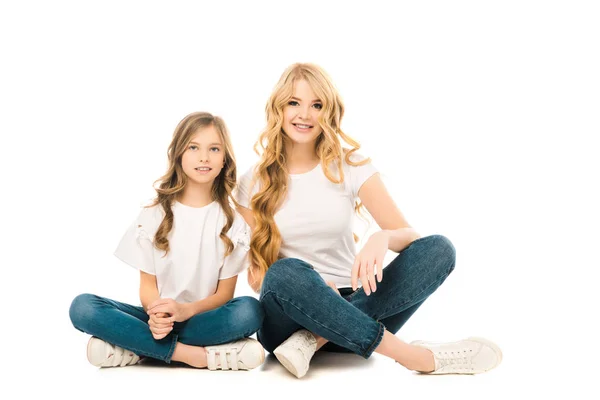 Sonrientes madre e hija sentadas en el suelo con las piernas cruzadas y mirando a la cámara sobre fondo blanco - foto de stock