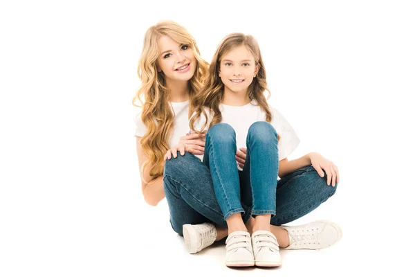 Smiling mother and daughter sitting on floor and looking at camera on white background — Stock Photo