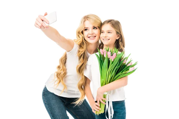 Sonriente madre tomando selfie con hija sosteniendo ramo de tulipanes aislados en blanco - foto de stock