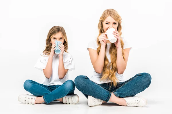 Madre e hija sentadas en el suelo con las piernas cruzadas y tomando café sobre fondo blanco - foto de stock