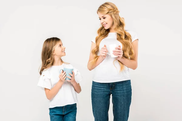 Sonrientes madre e hija sosteniendo copas y mirándose el uno al otro sobre fondo blanco - foto de stock