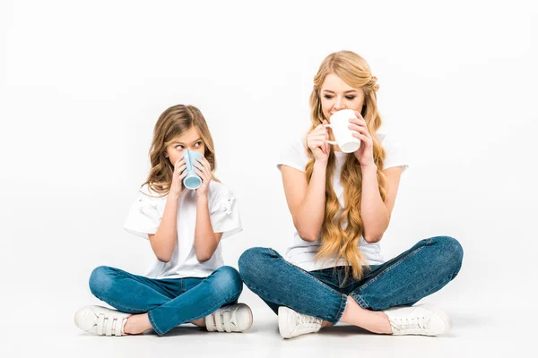 Cheerful mother and daughter drinking coffee while sitting on floor with crossed legs on white background — Stock Photo