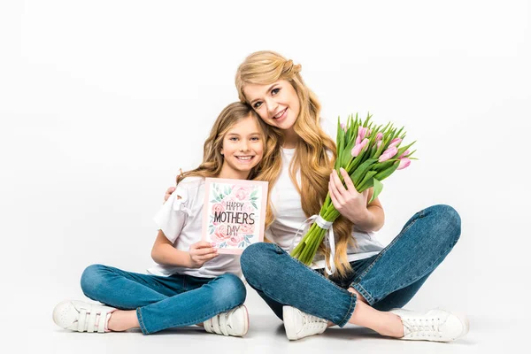 Madre con ramo de tulipanes rosados e hija con feliz tarjeta de felicitación del día de las madres sentada en el suelo con las piernas cruzadas sobre fondo blanco - foto de stock