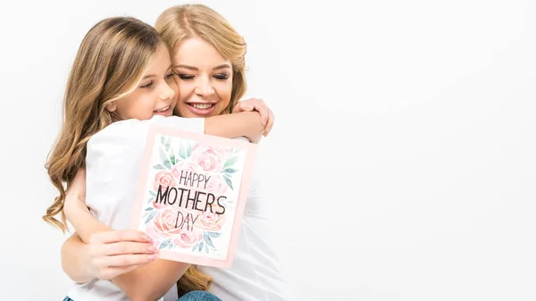 Очаровательный ребенок обнимает улыбающуюся мать с поздравительной открыткой на белом фоне — стоковое фото