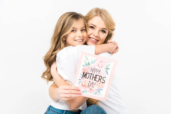 Feliz hermosa madre sosteniendo feliz madre día tarjeta de felicitación y abrazando adorable hija sobre fondo blanco - foto de stock