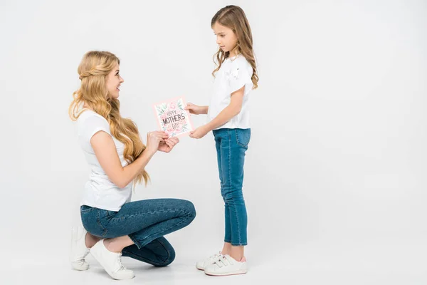 Hockende Mutter nimmt Glückwunschkarte zum Muttertag von lächelnder Tochter auf weißem Hintergrund — Stockfoto