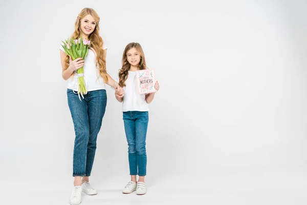 Lächelnde Mutter mit einem Strauß pinkfarbener Tulpen und lächelnde Tochter mit einer Glückwunschkarte zum Muttertag, die Händchen haltend in die Kamera auf weißem Hintergrund blickt — Stockfoto
