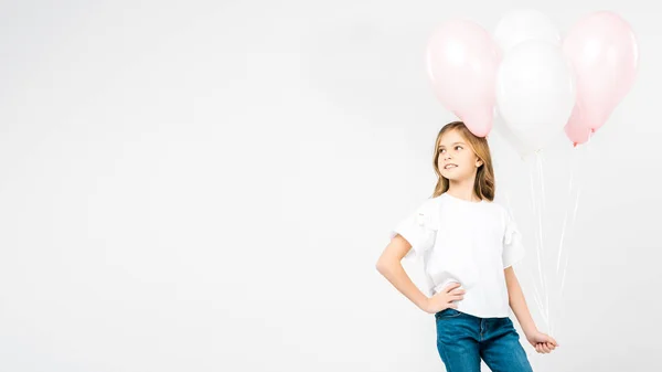 Criança sorridente com a mão no quadril segurando balões festivos de ar rosa e branco no fundo branco com espaço de cópia — Fotografia de Stock