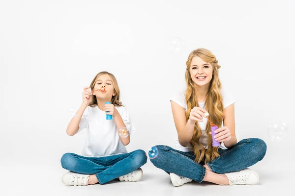 Cheerful mother and daughter sitting on floor with crossed legs and blowing soap bubbles on white background — Stock Photo